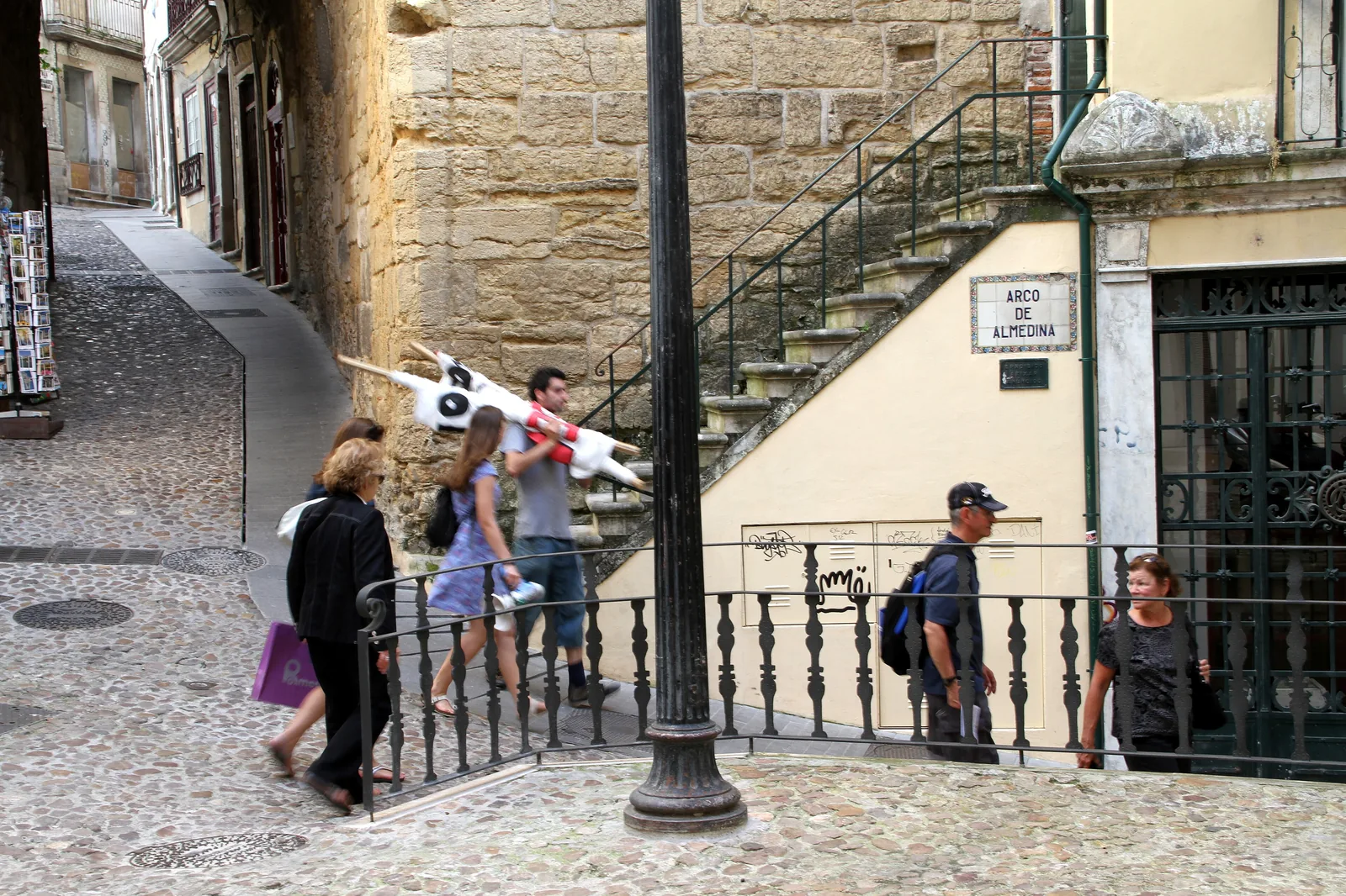 Visitors on the steps beside the Almedina arch, with a street lamp, an iron railing and the arch's name plate on the wall