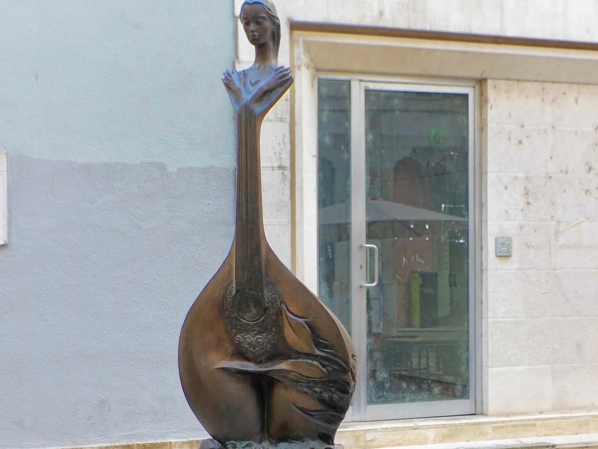 A bronze figure shaped like a Portuguese guitar stands on cobbles in front of a pale wall