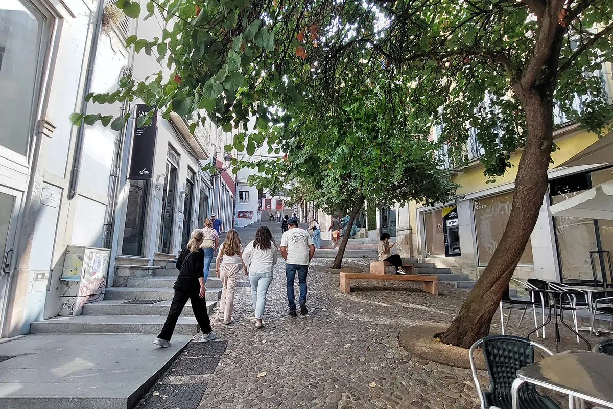 A paved street with a leafy tree, a flight of steps and visitors walking up between pale houses on Rua de Quebra-Costas