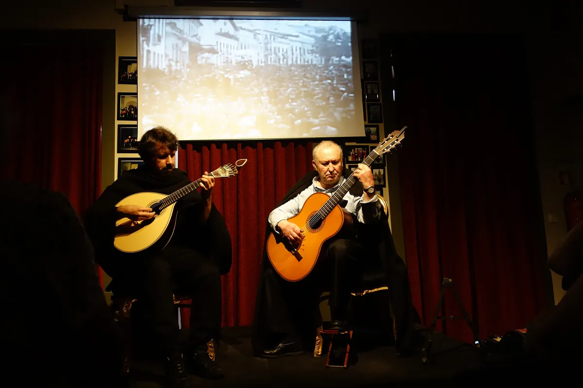 Two musicians in black capes sit on a small stage, one with a Coimbra guitar and one with a classical guitar, in front of red curtains and a projection screen