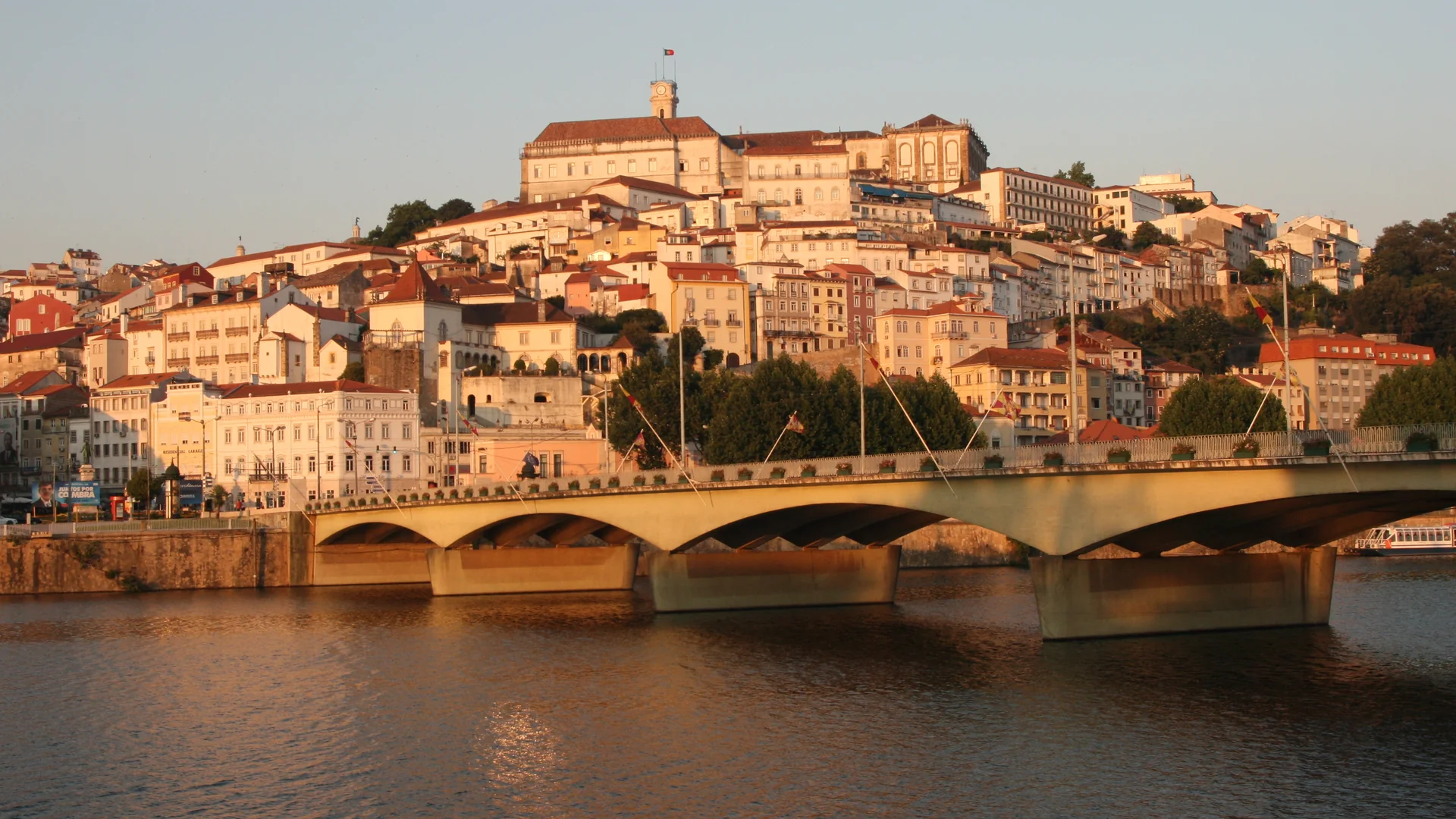 Coimbra's old town climbing a hill above a bridge over the Mondego River, its pale houses and red roofs lit gold by the evening sun