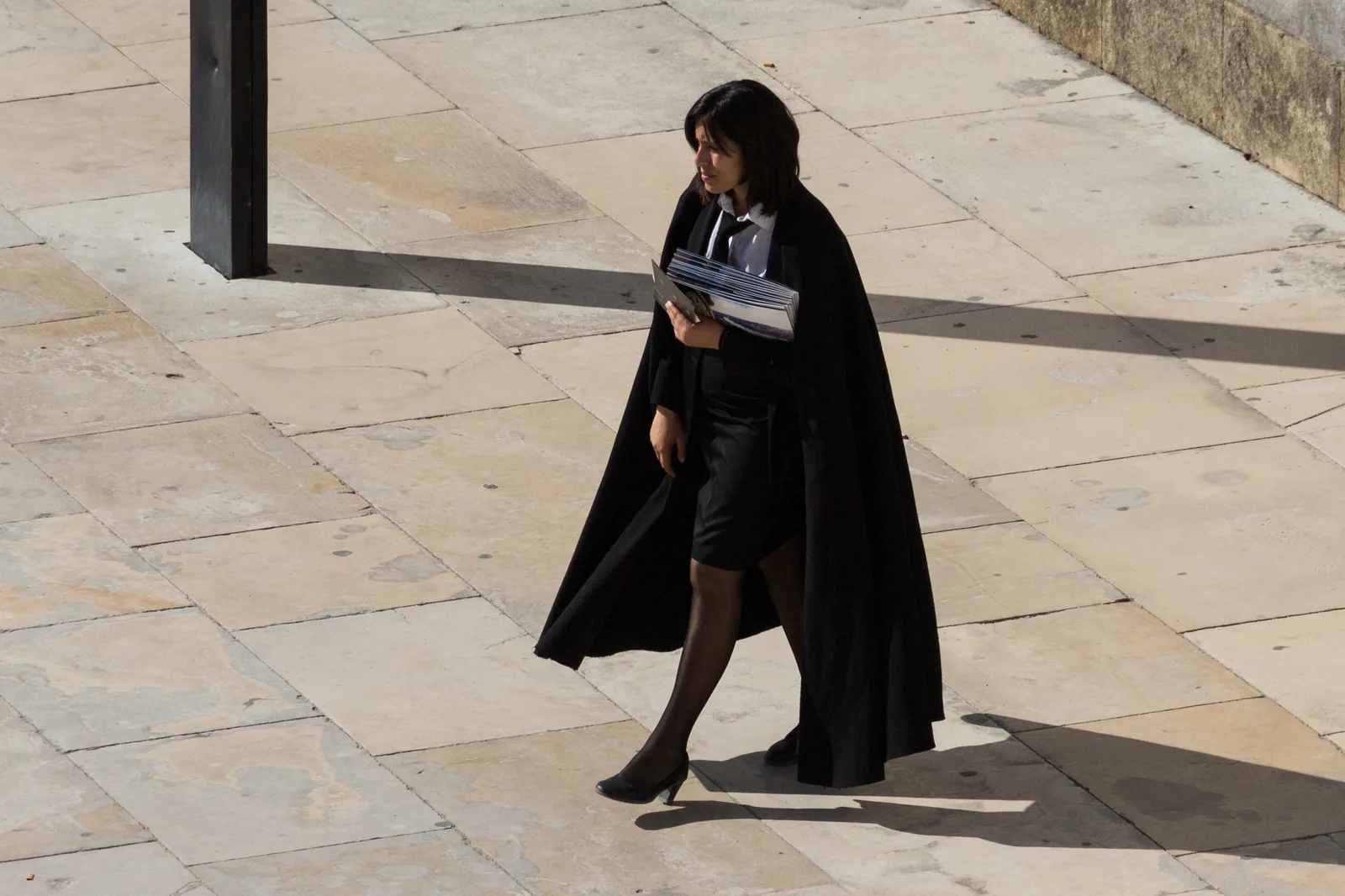 A student in a black academic cape walks across a paved square with a folder in her hand